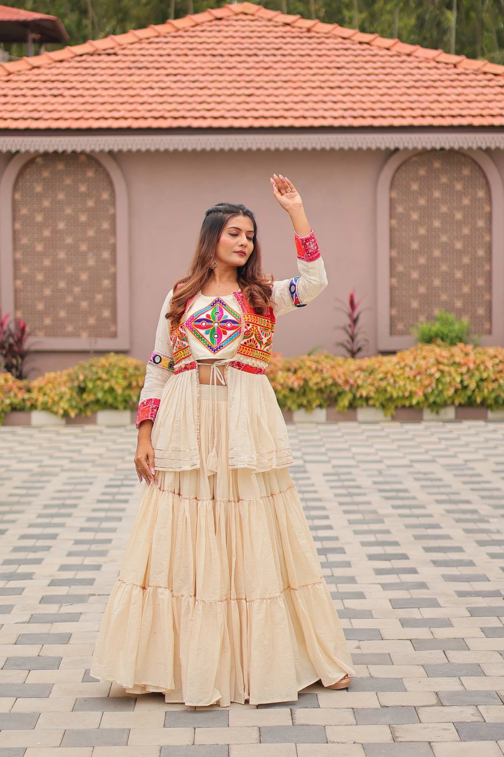 Woman in a traditional outfit standing on a paved area with a building and flowers in the background